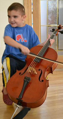 A photograph of a small child in crocs and a Nordiques t-shirt playing a cello.
